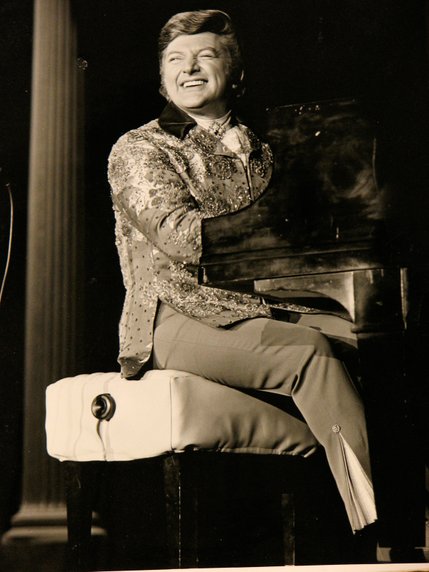 Liberace winks at the audience while playing his grand piano.  His big smile, infectious enthusiasm and flamboyant sequined outfits earned him the nickname 