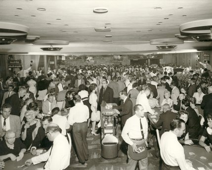 Guests crowd the table games pit at the Sands Hotel's main gaming area in 1959. On the left and right sides, men can be seen holding flash bulbs to illuminate the back of the normally darkened room. Guests often wore their finest attire for a night of gambling and drinking in Las Vegas.