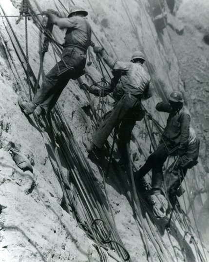 In April 1933, high scalers work to drill holes that contain dynamite on the rock wall on the Nevada side of Black Canyon. The work was dangerous, hard labor, often in temperatures above 100-degree heat. Workers earned about 50 cents an hour.