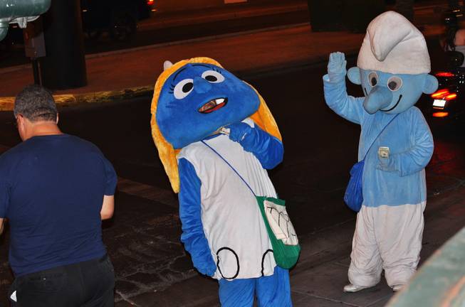 A performer's mustache is seen underneath his Smurfette costume as he leans back to scratch his neck while another performer in a smurf costume is seen greeting a passerby while holding his money September 2012.