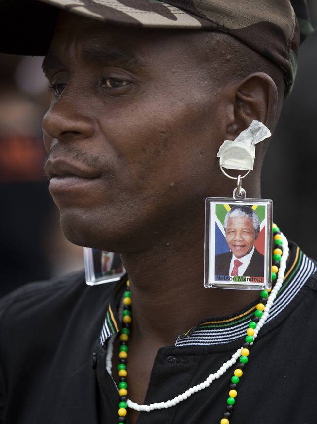A man wears key rings showing the face of Nelson Mandela taped to his ears to mimic earrings as he and others celebrate his life, in the street outside his old house in Soweto, Johannesburg, South Africa, Friday, Dec. 6, 2013. 
