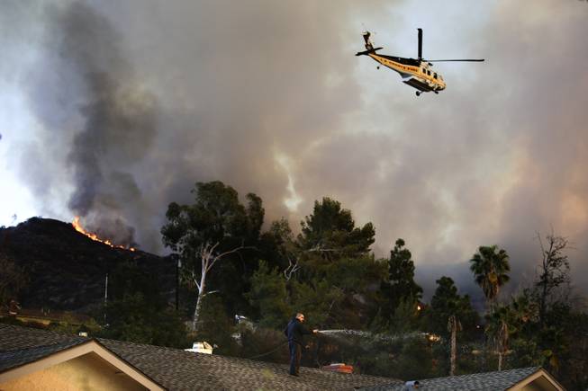 A helicopter carrying water flies over the residential area as a man sprays water on his home on Thursday, Jan. 16, 2014, in Azusa, Calif. 