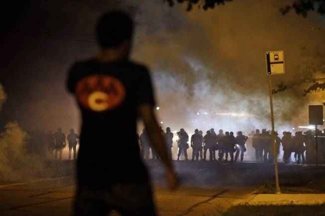 AP10ThingsToSee - A man watches as police walk through a cloud of smoke during a clash with protesters Wednesday, Aug. 13, 2014, in Ferguson, Mo. Protests in the St. Louis suburb rocked by racial unrest since a white police officer shot an unarmed black teenager to death turned violent Wednesday night, with people lobbing Molotov cocktails at police who responded with smoke bombs and tear gas to disperse the crowd.