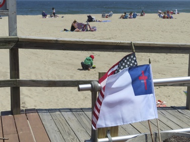 People sit on the beach on a weekday afternoon in Ocean Grove, N.J. on May 2, 2024, as a Christian flag and an American flag flutter in the breeze. The state of New Jersey says the Ocean Grove Camp Meeting Association is violating state beach access laws by keeping people off the beach until noon on Sundays.
