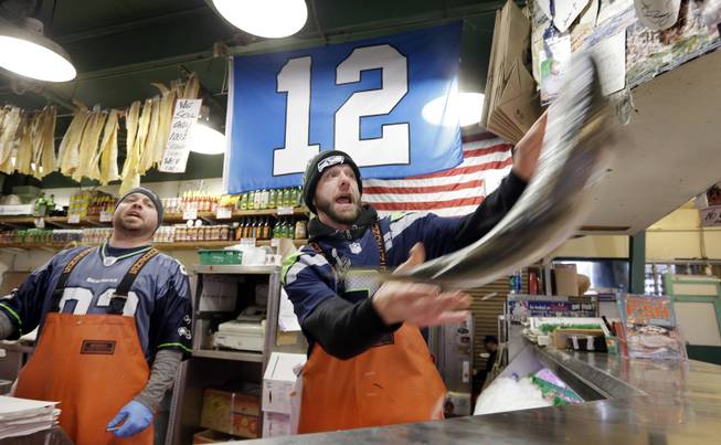 Fish monger Justin Hall, right, launches a salmon as Scott Smith looks on as the pair wear Seattle Seahawks' jerseys and work beneath a "12th Man" flag at the Pike Place Fish Market, Monday, Jan. 27, 2014, in Seattle. Seattle and Denver are both awash in team colors and symbols as Super Bowl Sunday approaches, when the Seahawks play the Denver Broncos in New Jersey for the NFL championship.