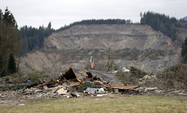 A flag, put up by volunteers helping search the area, stands in the ruins of a home left at the end of a deadly mudslide from the now-barren hillside seen about a mile behind, Tuesday, March 25, 2014, in Oso, Wash. At least 14 people were killed in the 1-square-mile slide that hit in a rural area about 55 miles northeast of Seattle on Saturday. Several people also were critically injured, and homes were destroyed.