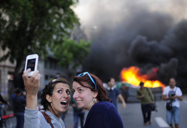 Tourists take a 'selfie' picture as demonstrators burn a trash container during a May Day rally in Barcelona, Spain, Thursday, May 1, 2014. Tens of thousands of workers marked May Day in European cities with a mix of anger and gloom over austerity measures imposed by leaders trying to contain the eurozone's intractable debt crisis.